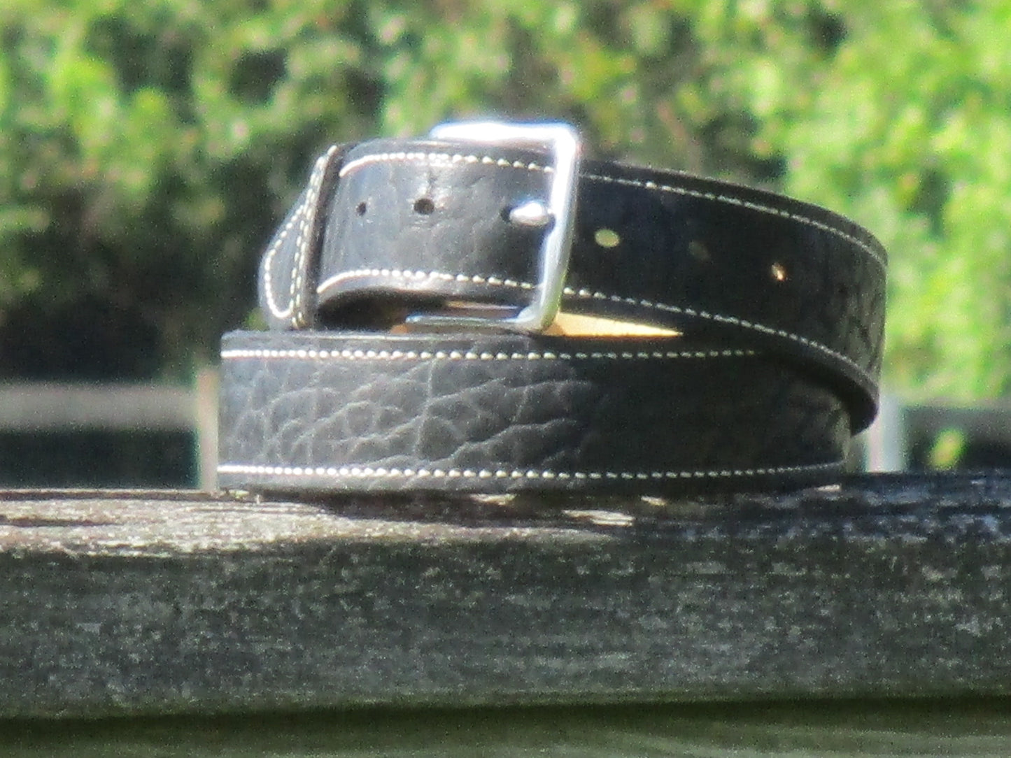 North American Bison Black leather belt with a full grain leather, contrast stitched liner, with a Stainless Steell buckle on a wooden surface with a blurred green background
