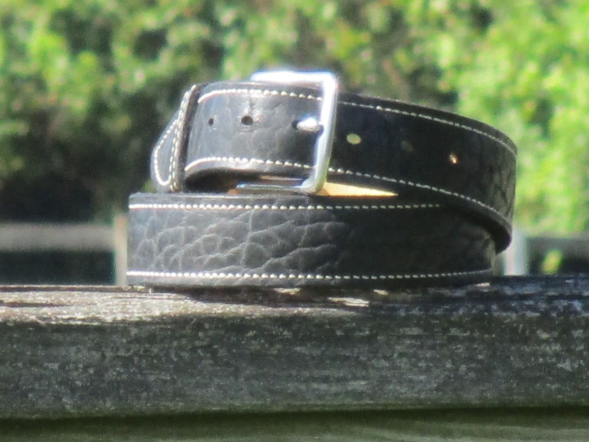 North American Bison Black leather belt with a full grain leather, contrast stitched liner, with a Stainless Steell buckle on a wooden surface with a blurred green background