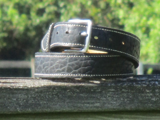 North American Bison Black leather belt with a full grain leather, contrast stitched liner, with a Stainless Steell buckle on a wooden surface with a blurred green background