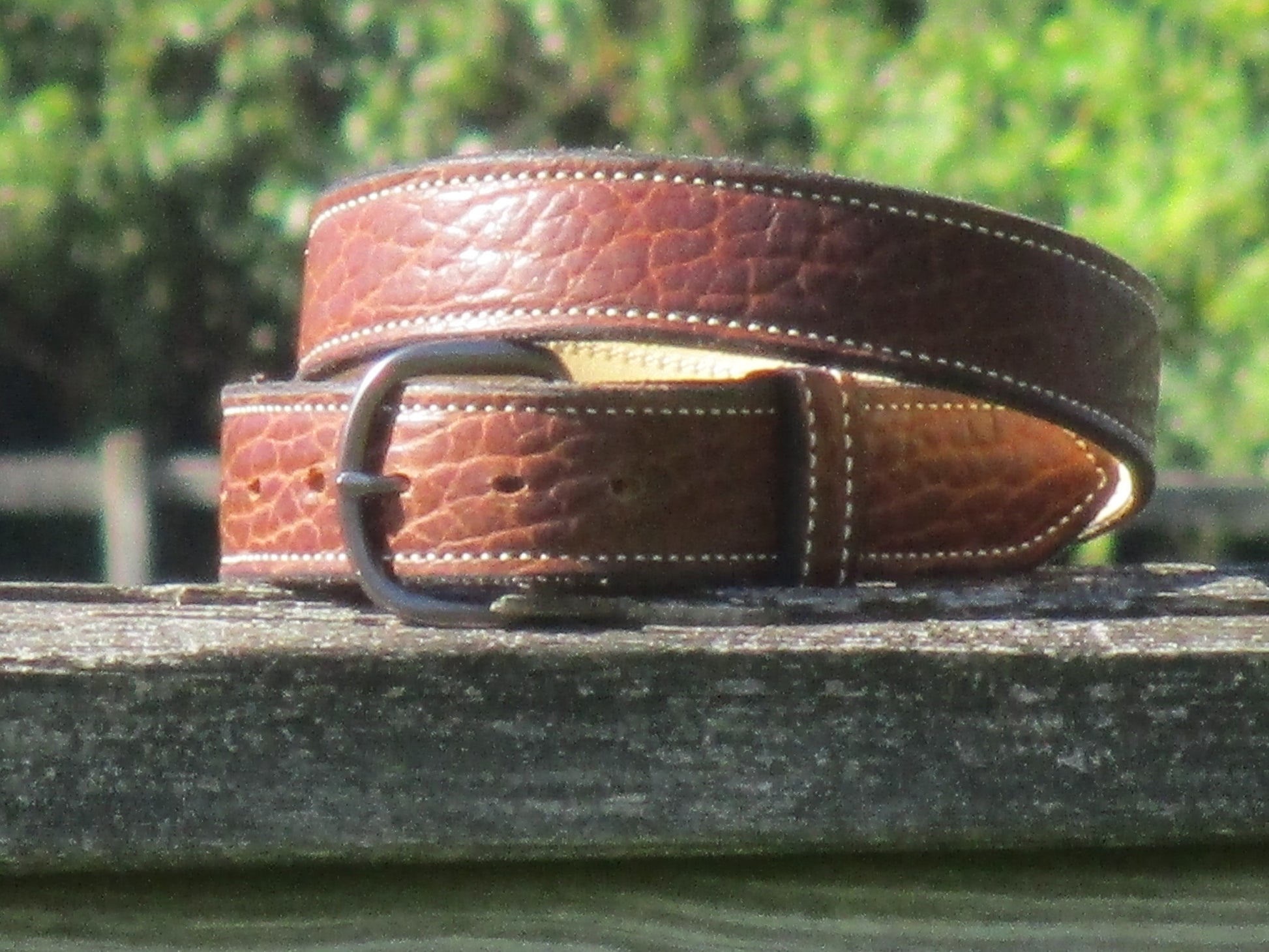 North American Bison Brown leather belt with a full grain leather, contrast stitched liner, with a Gun Metal color metal buckle on a wooden surface with a blurred green background