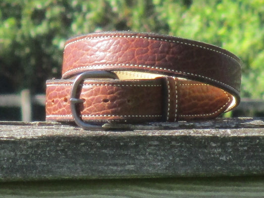 North American Bison Brown leather belt with a full grain leather, contrast stitched liner, with a Gun Metal color metal buckle on a wooden surface with a blurred green background