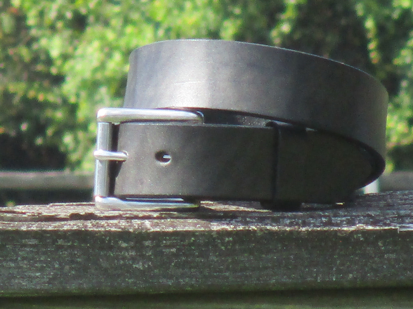 Black leather belt made from 12/13 oz English Bridle Leather with Stainless Steel buckle on a wooden surface with a blurred green background