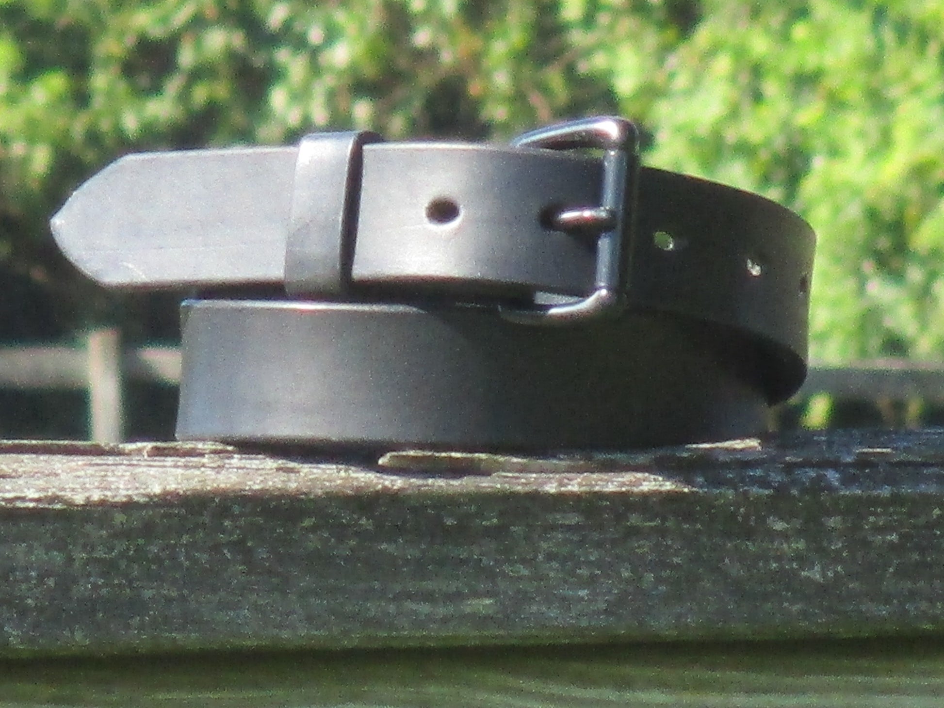 Black leather belt made from 9/10 oz. English Bridle Leather with a Black metal buckle on a wooden surface with a blurred green background