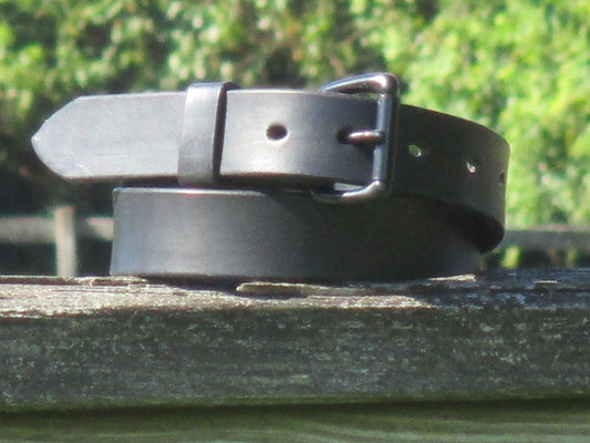 Black leather belt made from 9/10 oz. English Bridle Leather with a Black metal buckle on a wooden surface with a blurred green background