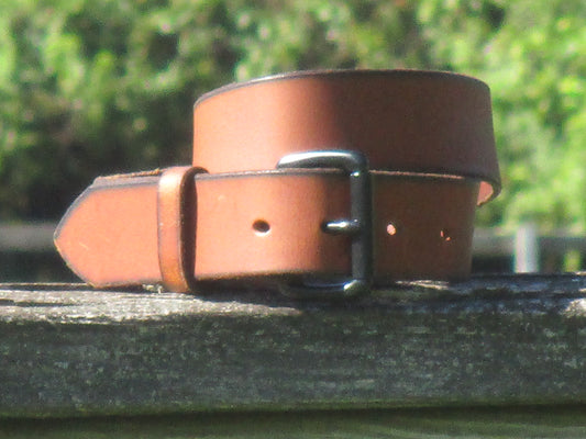 Water Buffalo Medium Brown  leather belt with Black metal buckle on a wood surface with a blurred green background.